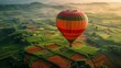 A hot air balloon is flying over a field of crops. The balloon is orange and green. The sky is cloudy and the sun is setting