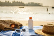 Sun protection gear laid out on seaside day. Straw hat, sunglasses, sunscreen bottle on blue towel. Summer, holiday essentials on sandy beach, against ocean backdrop. Skin care products in sunlight.