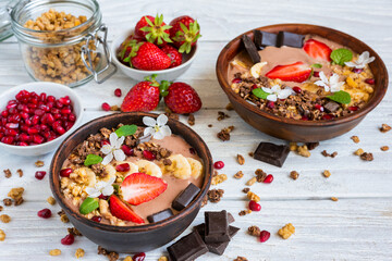 Wall Mural - Chocolate smoothie bowl topped with oat granola, banana, strawberries, pomegranate and spring flowers on white wooden table. Healthy vegan protein food with cocoa for breakfast