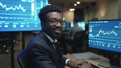 Attractive African American broker with elegant suit using computer for business trading. Young man cheerfully smiling at camera while working on crypto charts. On displays visible financial graphs.