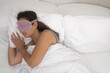 A picture of peacefulness, a woman in her sleep mask sleeps soundly on a pillow beneath a comforting blanket, a testament to healthy sleep habits. 