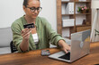 A woman in spectacles wields a credit card beside her laptop, efficiently handling online payments and shopping. 
