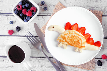 Sticker - Fun child theme breakfast pancake in the shape of a dinosaur. Top view table scene on a white wood background.