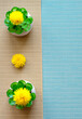 three yellow dandelions and two green artificial decors on a brown and blue striped tablecloth. Free place for text and logo
