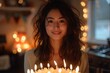 A young woman with a warm smile, holding a birthday cake adorned with glowing candles in a cozy ambiance