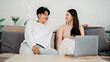 © Satori Studio - Young couple in casual attire enjoys a comfortable moment on their couch with a laptop, sharing a conversation in a modern living space.