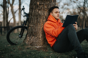 Casual young man enjoys downtime in the park, sitting by a tree with his notebook, mountain bike standing in the background.