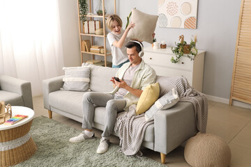 Wall Mural - Young man in headphones using mobile phone and his naughty little son with pillow at home