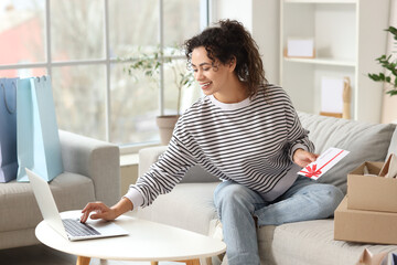 Poster - Young African-American woman with gift card and laptop shopping online on sofa at home