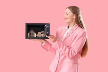 Wall Mural - Beautiful young happy woman with safe box, piggy bank and model of house on pink background