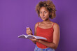 Young smiling African American woman student holding workbook and pen doing instructions from teacher to prepare for exam to receive cash scholarship stands in purple studio. Education, knowledge