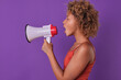 Young curly excited African American woman promoter holds white megaphone and loudly announces advertising information fulfilling instructions of marketer to attract buyers stands in purple studio.