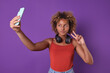Young optimistic casual African American woman blogger broadcasts video via phone and makes V hand gesture welcoming new subscribers or thanking viewers for donating posing on purple background.