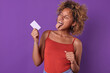 Young beautiful cheerful African American woman with credit card in hands shows tongue rejoicing at opportunity to use borrowed funds from bank to make non-cash payments stands in purple studio.