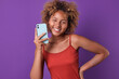 Young laughing ethnic African American woman teen with smartphone holds hand on belt and looks at camera with wide smile feeling joy due to excellent coincidence stands on purple background.