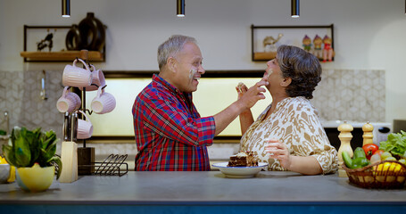 Wall Mural - Indian Asian Hindu older lover gen x couple pair sitting modern kitchen pastry cake cream put apply on face enjoy day indoor house happy old aged elder man male husband wife female make fun joy home