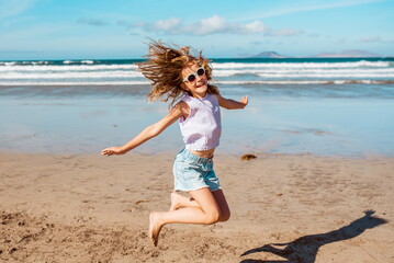 Jumping girl on beach. Smilling blonde girl enjoying sandy beach, looking at crystalline sea in Canary Islands. Concept of beach summer vacation with kids.