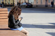 Girl in black leather jacket sitting with smartphone on а street bench in spring