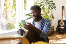 Smiling Non-binary Person Holding Coffee Cup And Using Smart Phone At Home