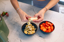 Woman Slicing Banana Next To Strawberry Bowl In Kitchen At Home