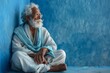 Elderly Indian Man in Traditional Clothing Reflecting While Seated Against Blue Wall