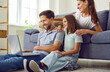 © Studio Romantic - Happy young family of three with child girl sitting on the floor at home using laptop together having video call talking online. Smiling parents with daughter watching funny cartoon or movie.