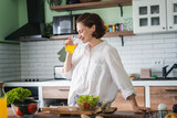 Fototapeta Zachód słońca - Young caucasian smiling woman drinking orange juice while standing in the kitchen at home, vitamins and healthy eating