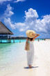 A elegant holiday woman in a white dress and hat walks down a tropical paradise beach in the Maldives islands