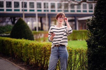 Wall Mural - In a city park, a young, trendy woman adjusts her glasses, phone in hand, epitomizing urban lifestyle and the integration of nature within metropolitan areas.