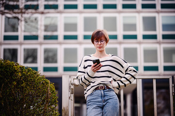 Wall Mural - A contemplative woman stands by the railing, smartphone in hand, against the architectural backdrop of a cityscape, blending leisure with the urban environment.