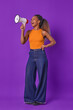 Young positive beautiful African American woman with white megaphone in hands works as promoter advertising new shopping center with excellent prices on goods stands on isolated purple background.