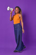 Young successful smiling African American business woman with megaphone in hand smiles turning head to camera and announcing launch of startup dressed in casual style stands posing in purple studio.