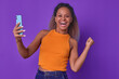 Young overjoyed inspired African American woman makes victorious wave hand and holds phone in hand after winning cash prize in lottery or casino stands in purple studio. Success, winning, achievement