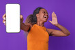Young happy ethnic African American woman student holds out mobile phone with white screen to camera and puts hand to mouth shouting advertising slogan posing on isolated purple background.