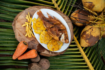 Wall Mural - Top view of a cooked fish and fried plantains on a log board on a palm tree with ingredients