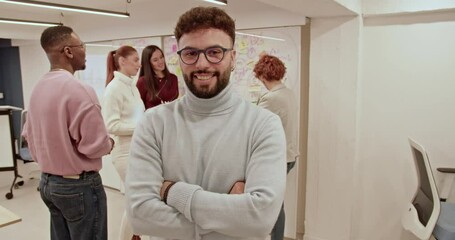 Wall Mural - A smiling business employee poses with a confident posture while colleagues discuss strategy on a whiteboard in a modern office, showcasing a positive and collaborative work environment.