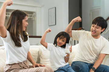 Wall Mural - Happy Asian family, mother and father cheering little 6 year old daughter drinking milk from glass, raised arm muscles and fist for good health, sitting on sofa at home. Selective focus on child