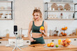 Attractive fitness blogger with blond hair recording video on smartphone about healthy dieting and food. Young woman in green sport outfit standing on kitchen and holding glass of fresh orange juice.