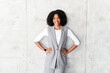 A poised African-American businesswoman stands with hands on her hips, wearing a stylish grey sleeveless vest and trousers, radiating confidence and professionalism against a textured grey background.