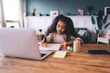 © BullRun - Engaged African American girl, 6-8 years old, diligently drawing at a wooden table, with a laptop, smartphone, and stationery around her. She's wearing a soft beige sweater, concentrating on studying