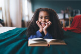 Fototapeta Nowy Jork - Joyful African American girl, aged 6-8, lying on her stomach on a green blanket, hands on cheeks, with a book open in front of her. Her bright smile and sparkling eyes, paired with a blue top