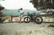 © Taras Grebinets - Handsome muscular man cyclist drinks water from sports bottle, standing by wooden fence next to electric mountain bike, on rural road overlooking white houses with red tile roofs in medieval village