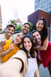 Fototapeta Londyn - Young group of people having fun enjoying free time together taking selfie portrait at city street. Millennial student friends laughing outdoors. Youth community concept.