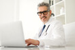 Senior doctor with grey hair looks at camera while using a laptop, suggesting adaptability to modern medical technology, friendly expression and attentive posture convey a commitment to patient care