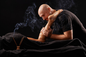 Wall Mural - handsome male masseur doing a massage on a girl's leg on a black background, concept of therapeutic relaxing massage