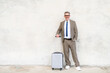 With a bright smile and a casual stance, a senior experienced businessman stands with his suitcase against concrete wall, embodying the ease and comfort of contemporary business travel