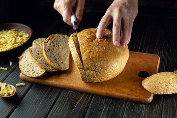 Canvas Print - Male hands with a knife cut fresh rye bread on a kitchen board before serving for lunch. Low key concept of healthy eating or baked goods