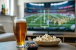 A glass of beer sits on a table next to a bowl of popcorn and a remote control, with a large television screen displaying a football game