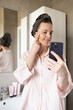 Woman smiling, applying cosmetic cream and looking at smartphone in bathroom, vlogging. Blogger recording morning skincare routine, user generated content