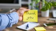 Close-up of a hand presenting a yellow sticky note with Thank You on a blurry office desk background